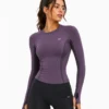 Model front 3-4 Female model wearing a fitted plum purple women’s technical activewear top with long sleeves, breathable mesh panels, contoured seams, thumbhole cuffs, and athletic styling