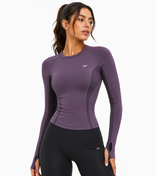 Model front 3-4 Female model wearing a fitted plum purple women’s technical activewear top with long sleeves, breathable mesh panels, contoured seams, thumbhole cuffs, and athletic styling