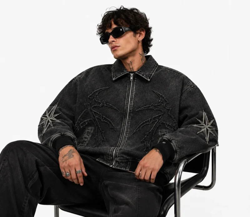men-wearing-custom-jean-jacket Men's custom jean jacket styled for streetwear, featuring black washed denim, front zipper closure, artistic embroidery, sleeve patches, ribbed waistband and cuffs.