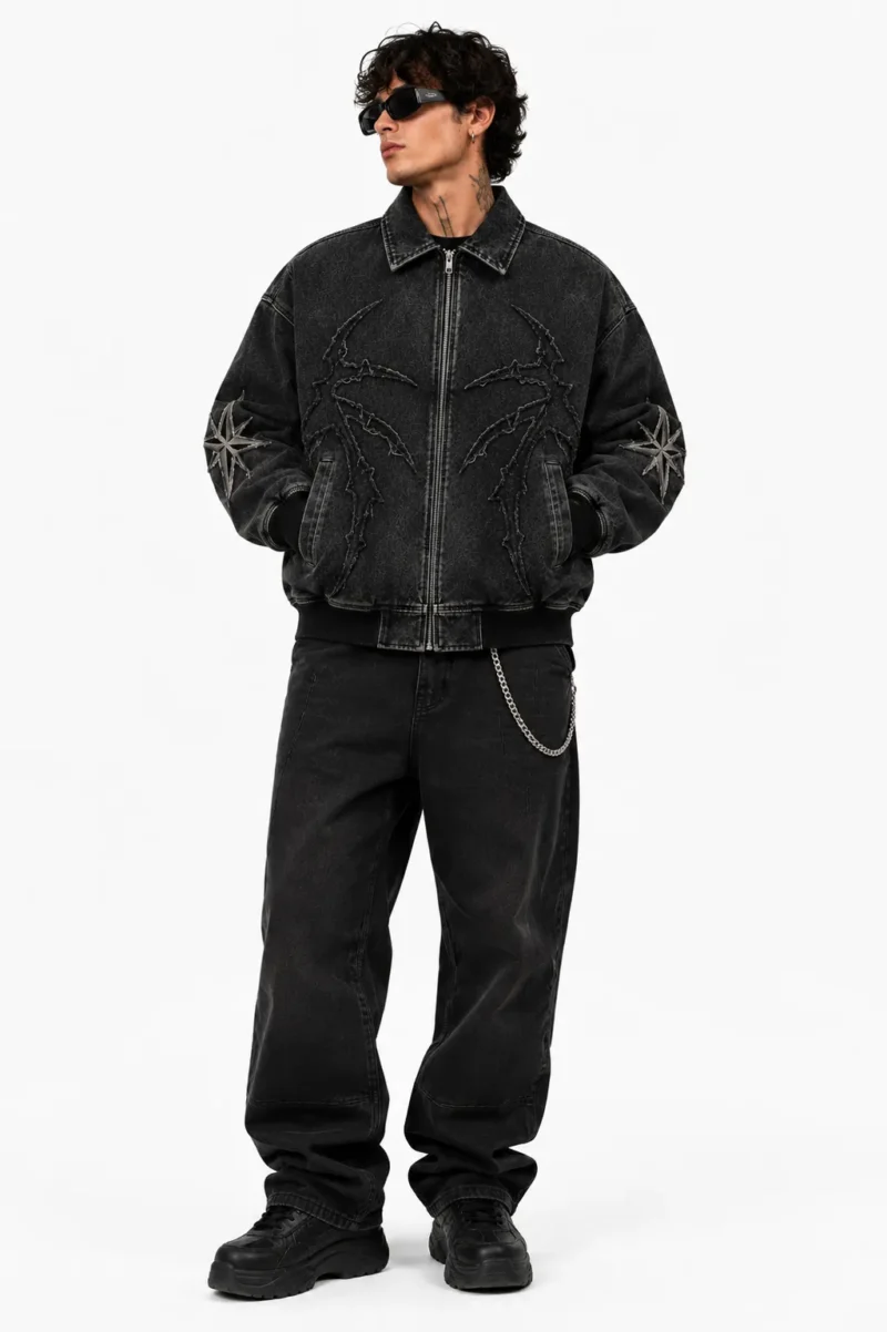 mens-custom-jean-jacket-streetwear Man wearing a custom jean jacket made from washed black denim, featuring a front zipper, embroidered artwork, decorative sleeve patches, ribbed cuffs and a modern streetwear fit.